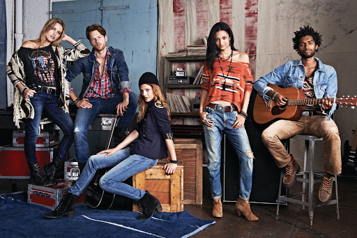 Group fashion shot in a studio with five people wearing denim jeans, jackets, and boots, posing against a backdrop with music equipment.
