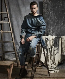 Man wearing a denim top and distressed jeans seated on a wooden ladder in a studio with painted backdrops.