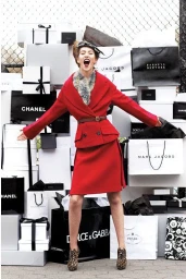 Smiling woman in a red skirt suit with a fur collar standing in front of a massive stack of luxury shopping bags
