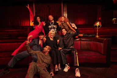 A group of seven diverse models posing together on a red leather booth in a dimly lit bar interior.