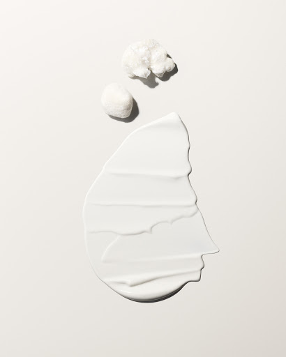 Close-up of a creamy white cosmetic skincare product swatch alongside two cotton pads against a neutral background with soft lighting.