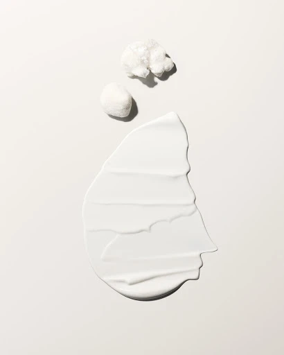 Close-up of a creamy white cosmetic skincare product swatch alongside two cotton pads against a neutral background with soft lighting.