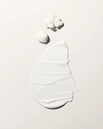 Close-up of a creamy white cosmetic skincare product swatch alongside two cotton pads against a neutral background with soft lighting.