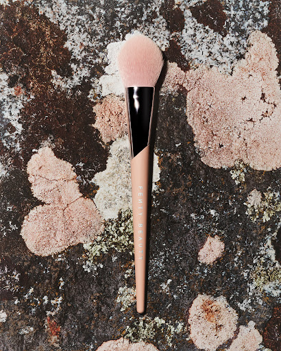 Beauty product shot: A Fenty Beauty powder brush lies on a lichen-covered rock in natural light, highlighting its soft, pink bristles.