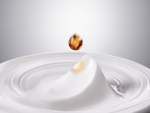 Close-up of a falling amber-colored drop of oil onto rippled white cream, creating a splash. Shot in a bright photo studio.