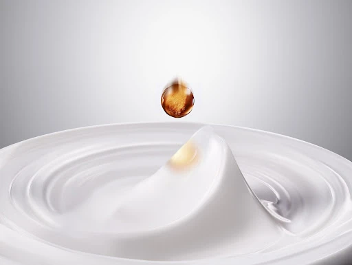 Close-up of a falling amber-colored drop of oil onto rippled white cream, creating a splash. Shot in a bright photo studio.