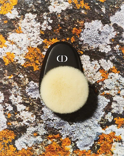 Overhead studio shot of a cosmetic powder puff with black applicator bearing the CD logo, placed on a textured, earthy surface.