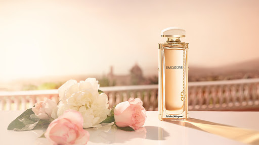 Elegant perfume bottle with gold accents next to pink and white flowers against a soft, blurred, sunny background on a white table.