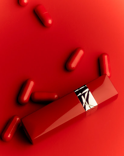 Red lipstick tube with silver accents, surrounded by scattered red capsules on a glossy red surface, product photography.