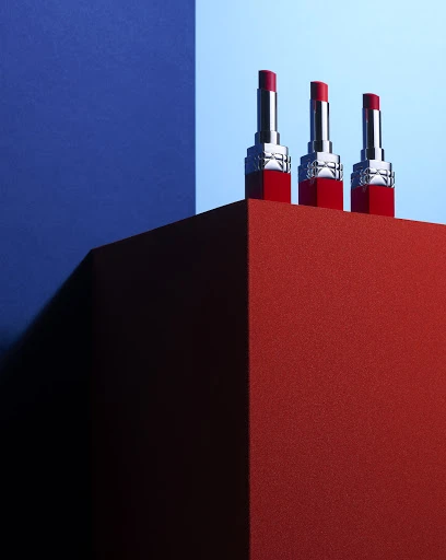 Three red lipsticks stand atop a red geometric surface against a dual-tone blue background, creating a high-contrast beauty product shot.