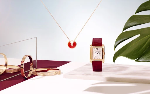 Gold watch with red strap, gold necklace with red pendant, and gold bangles arranged with a green leaf and acrylic panel