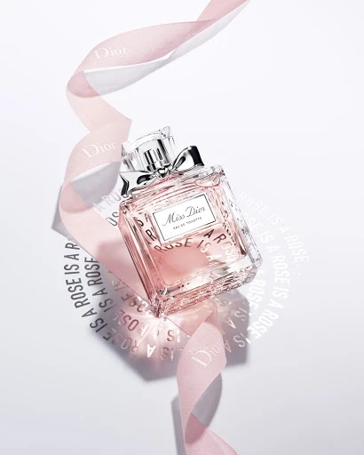 Miss Dior perfume bottle, adorned with a ribbon, sits on a white surface with shadow lettering; a beauty product shot for marketing.