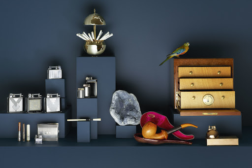 Still life arrangement featuring lighters, smoking pipes, a geode, and a bird figurine against a dark blue background.