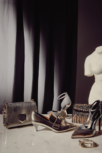 Fashion still life with gold and silver tone handbags, heels, and shoes arranged on a marble surface in front of a draped backdrop.