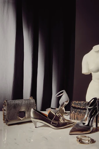 Fashion still life with gold and silver tone handbags, heels, and shoes arranged on a marble surface in front of a draped backdrop.