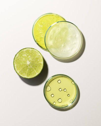Overhead shot of fresh lime half and three petri dishes filled with clear and green-tinted liquids on a white background.