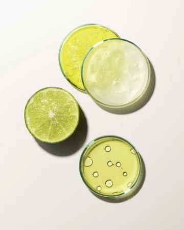 Overhead shot of fresh lime half and three petri dishes filled with clear and green-tinted liquids on a white background.