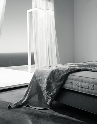 Monochromatic interior of bedroom with a bed covered by grey linen, white curtains and sea view from the terrace door in background.