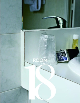 Bathroom counter scene: a transparent plastic cup, hotel amenities, and a watch, all reflected in a mirror above the sink.