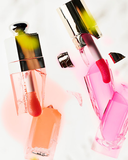 Still life shot of two broken cosmetic containers with metallic lids amidst spilled liquid foundation and lipstick in a light color palette.