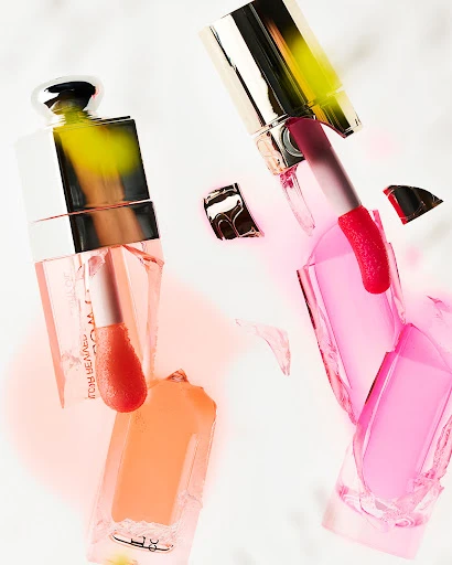 Still life shot of two broken cosmetic containers with metallic lids amidst spilled liquid foundation and lipstick in a light color palette.
