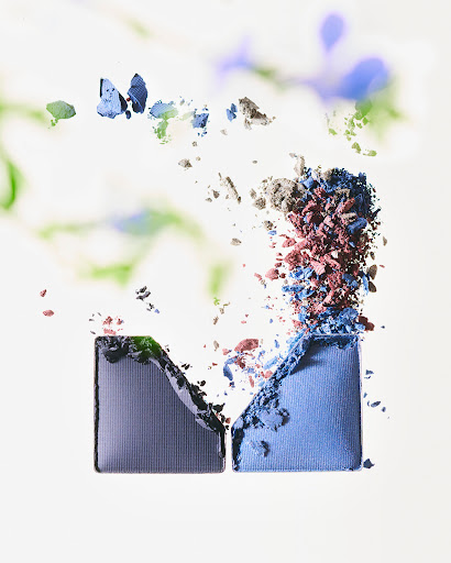 Cosmetic image featuring two eye shadow cases with powdered make-up spilling out in blue, brown, and gray against a blurred floral backdrop.