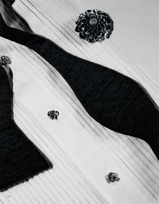 Monochrome studio still life of a formal white shirt, black bow tie, floral button covers, and a black flower on a white surface.