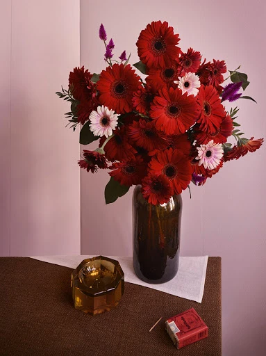Still life photograph with a dark glass vase filled with red gerbera daisies, an amber glass ashtray, and a red cigarette pack on a brown surface.