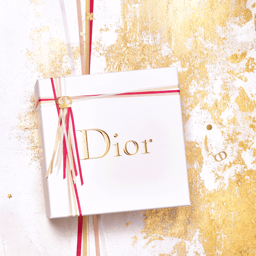 Elevated view of a white Dior gift box tied with red, gold and white ribbons and adorned with the Dior logo, set against a gold-splattered backdrop.