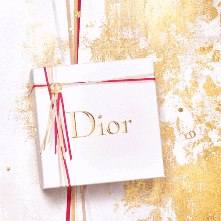 Elevated view of a white Dior gift box tied with red, gold and white ribbons and adorned with the Dior logo, set against a gold-splattered backdrop.