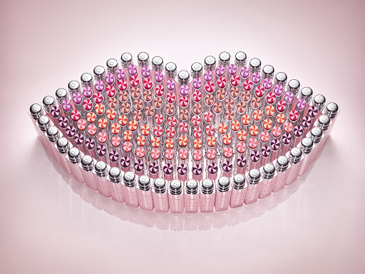 Arrangement of numerous lipstick tubes forming the shape of a pair of lips against a soft pink gradient background.