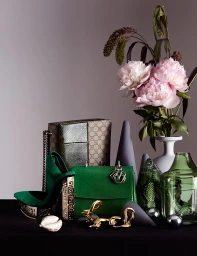 Still life arrangement with green handbags, a patterned shoulder bag, pink peonies in a white vase, and sculptural cones on a dark surface.