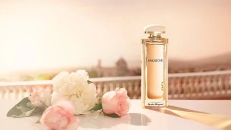 A perfume bottle and fresh white and pink roses on a light surface with a soft sunlit terrace background.
