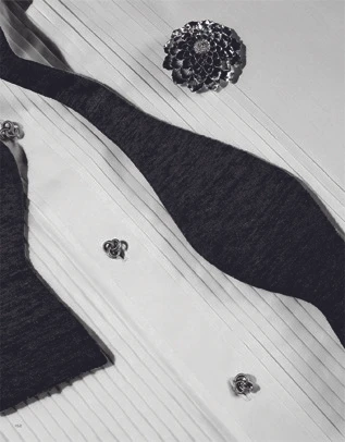 Close-up still life of a pleated white dress shirt with a black bow tie, metallic buttons, and a flower brooch.