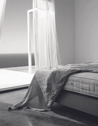 Black and white bedroom interior with a made bed, draped sheer curtains, and an open doorway overlooking the sea.