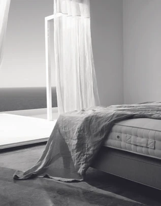 Black and white bedroom interior with a made bed, draped sheer curtains, and an open doorway overlooking the sea.