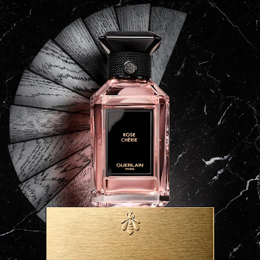Square product shot of a perfume bottle labelled Rose Cherie on a gold base against a black marble background with radiating wooden slats.