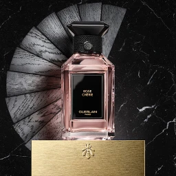 Square product shot of a perfume bottle labelled Rose Cherie on a gold base against a black marble background with radiating wooden slats.