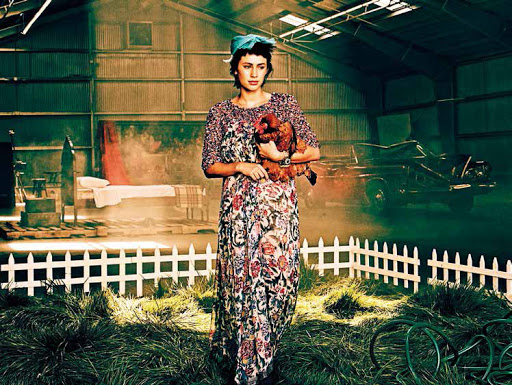 A woman in a floral dress stands in an indoor farm setting, holding a chicken with vintage car, bed, and garden hose in background.