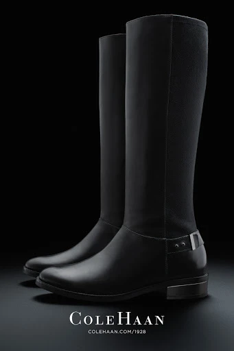 Studio shot of a pair of elegant black leather knee-high boots with dark background. Cole Haan logo and website are visible at the bottom.
