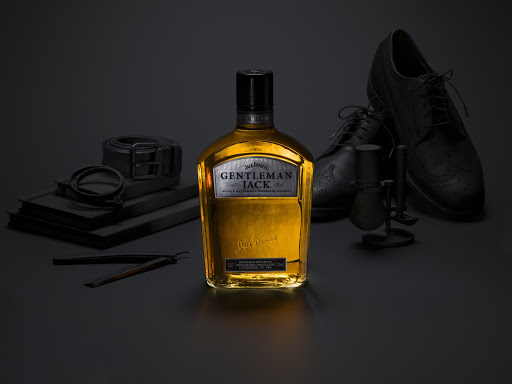 Studio shot of Jack Daniel's Gentleman Jack whiskey bottle with black leather dress shoes, shaving brush, and straight razor.