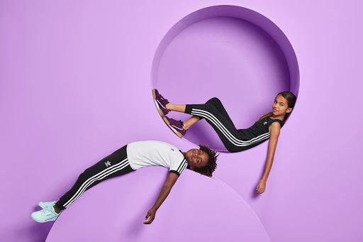 Two children pose in Adidas apparel against a lavender background, one inside a circular cutout and the other resting below it.