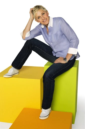 Adult female with short blond hair sitting on colorful cubes in a photo studio. She is wearing jeans and a light purple button-down shirt.