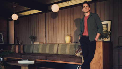 Adult male poses indoors in a green shirt and black pants. He is standing in front of a brown mid-century modern style wall.