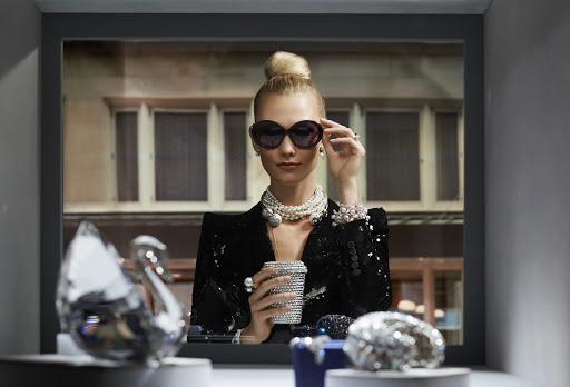 Elegant woman with sunglasses holds a jeweled cup in a display window of a shop with crystal figurines and pearl necklaces.