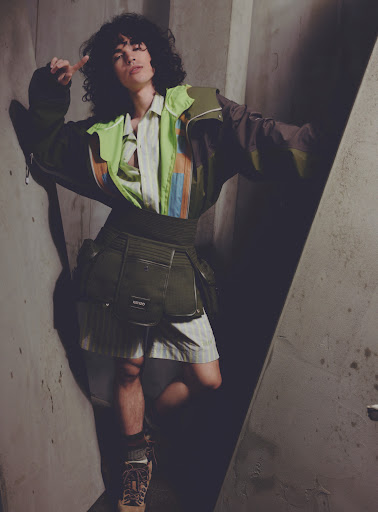 Fashion shot of a young adult with curly hair in an earth-toned outfit, standing in a concrete room with geometric light pattern.
