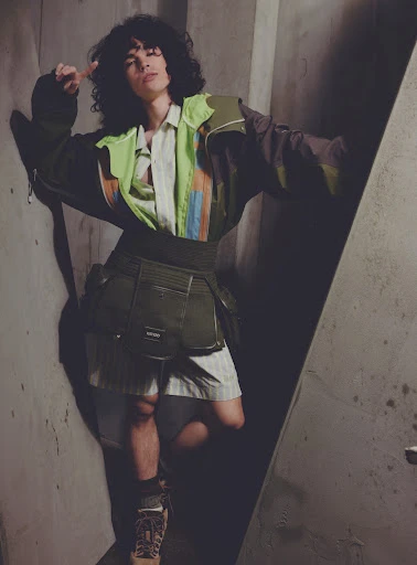 Fashion shot of a young adult with curly hair in an earth-toned outfit, standing in a concrete room with geometric light pattern.
