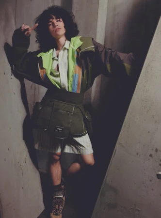 Fashion shot of a young adult with curly hair in an earth-toned outfit, standing in a concrete room with geometric light pattern.