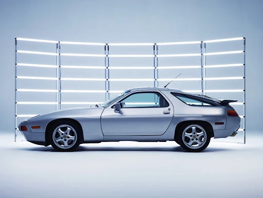Side view of a silver Porsche 928 sports car in a photo studio with a backdrop of white neon tube lights, clean product shot.