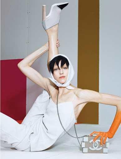 Woman in white outfit poses with high-heeled shoe and fashion purse in front of colorful graphic background in a studio setting.